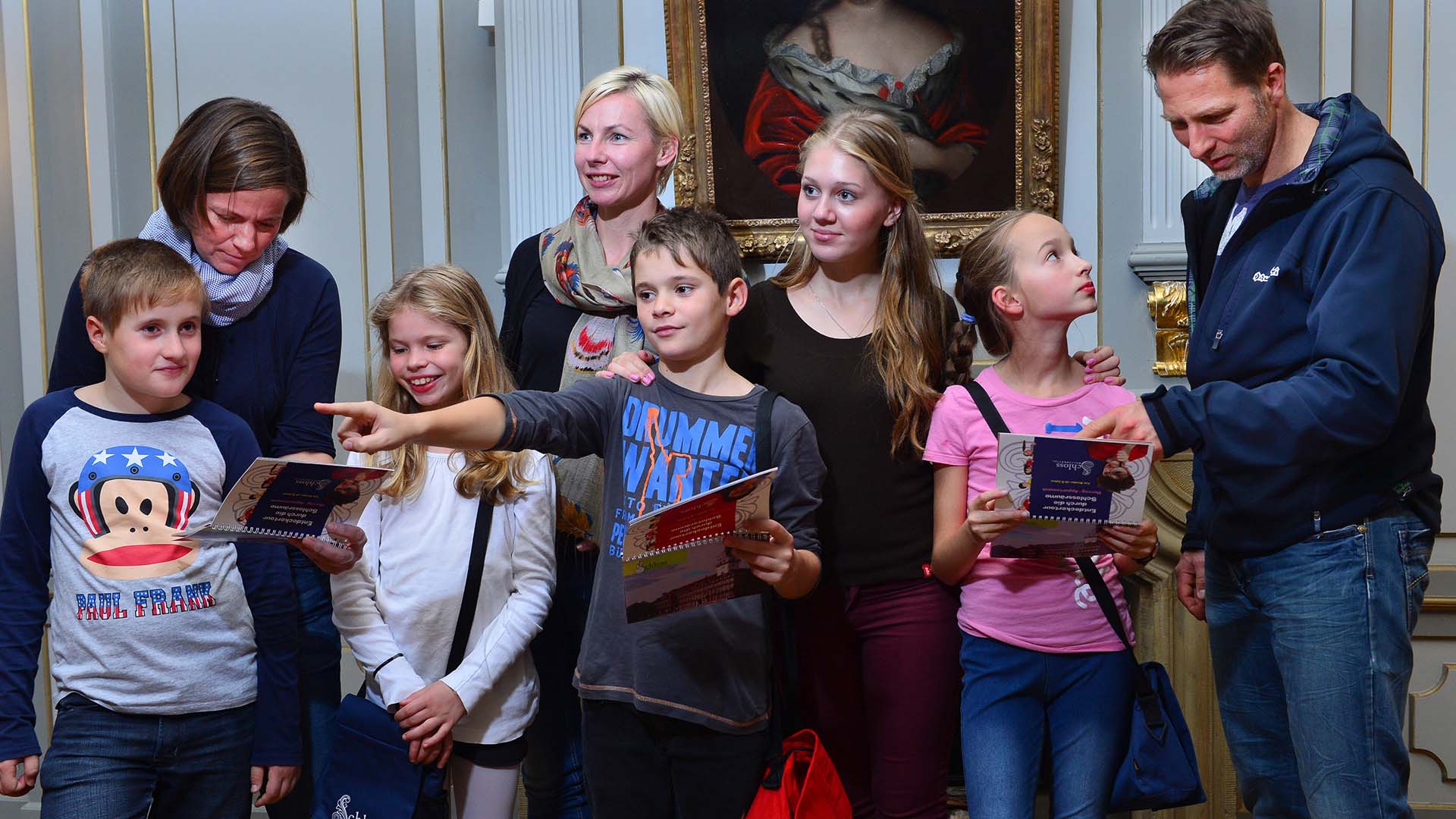 Das Foto zeigt Eltern mit ihren Kindern im Museum. Drei Erwachsene und fünf Kinder stehen in einem historischen Raum und schauen sich um. Drei der Kinder halten Hefte in den Händen. Ein Junge, mittig im Bild, zeigt auf etwas.