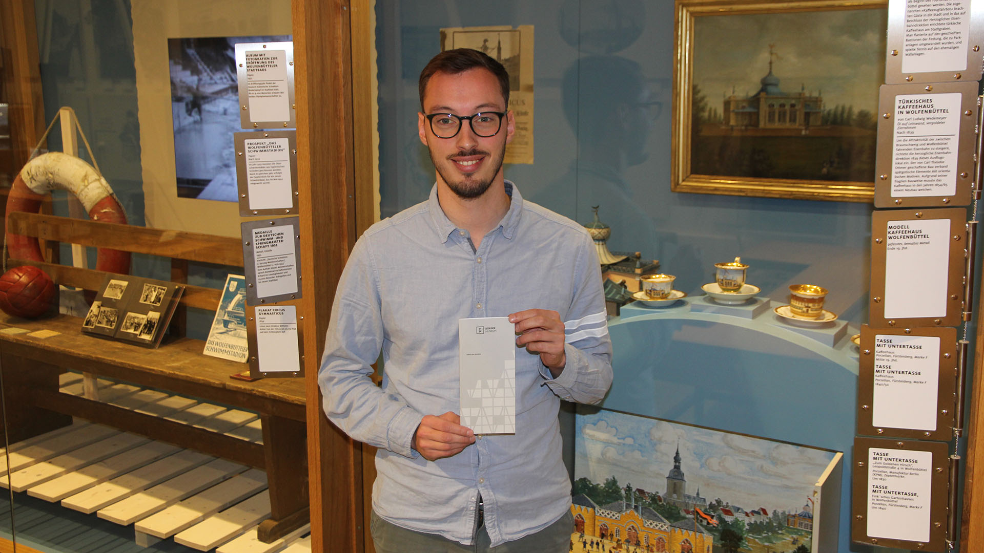 Vor einer großen Schauvitrine steht ein junger Mann und lächelt in die Kamera. In den Händen hält er den neuen« English Guide« des Bürger Museums.