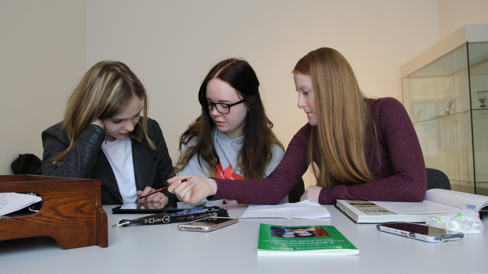 Drei ältere Schülerinnen sitzen vor einer Vitrine des Museums an einem Tisch und arbeiten zusammen an einem Tablet. Vor ihnen liegen Arbeitsmaterialien, Bücher und ein Exponat aus Holz.