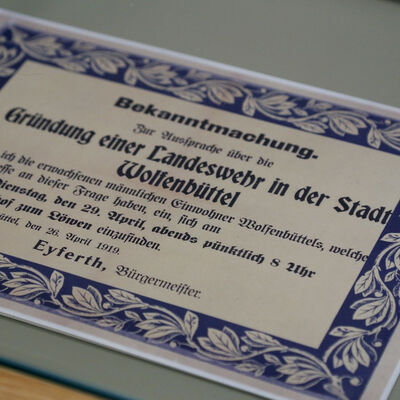 Auf dem Bild zu sehen ist unter einer Glasscheibe ein großes Plakat. Auf ihm steht ein Text zur Gründung einer Landwehr in Wolfenbüttel.