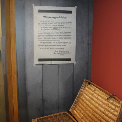 Das Foto zeigt die neugestaltetet Großvitrine im Bürger Museum. In ihr steht ein Korbwagen aus Bast, den Marie Deutschmann bei der Vertreibung hinter sich herzog. Die Wand dahinter ist rot, links befindet sich eine Fototapete mit einem aufgedruckten Plakat von 1946.