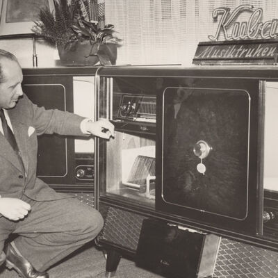 Schwarz-Weiß-Fotografie: Ein Mann in Anzug hockt vor einem großen Möbel mit dem Schriftzug »Kuba Musiktruhe«