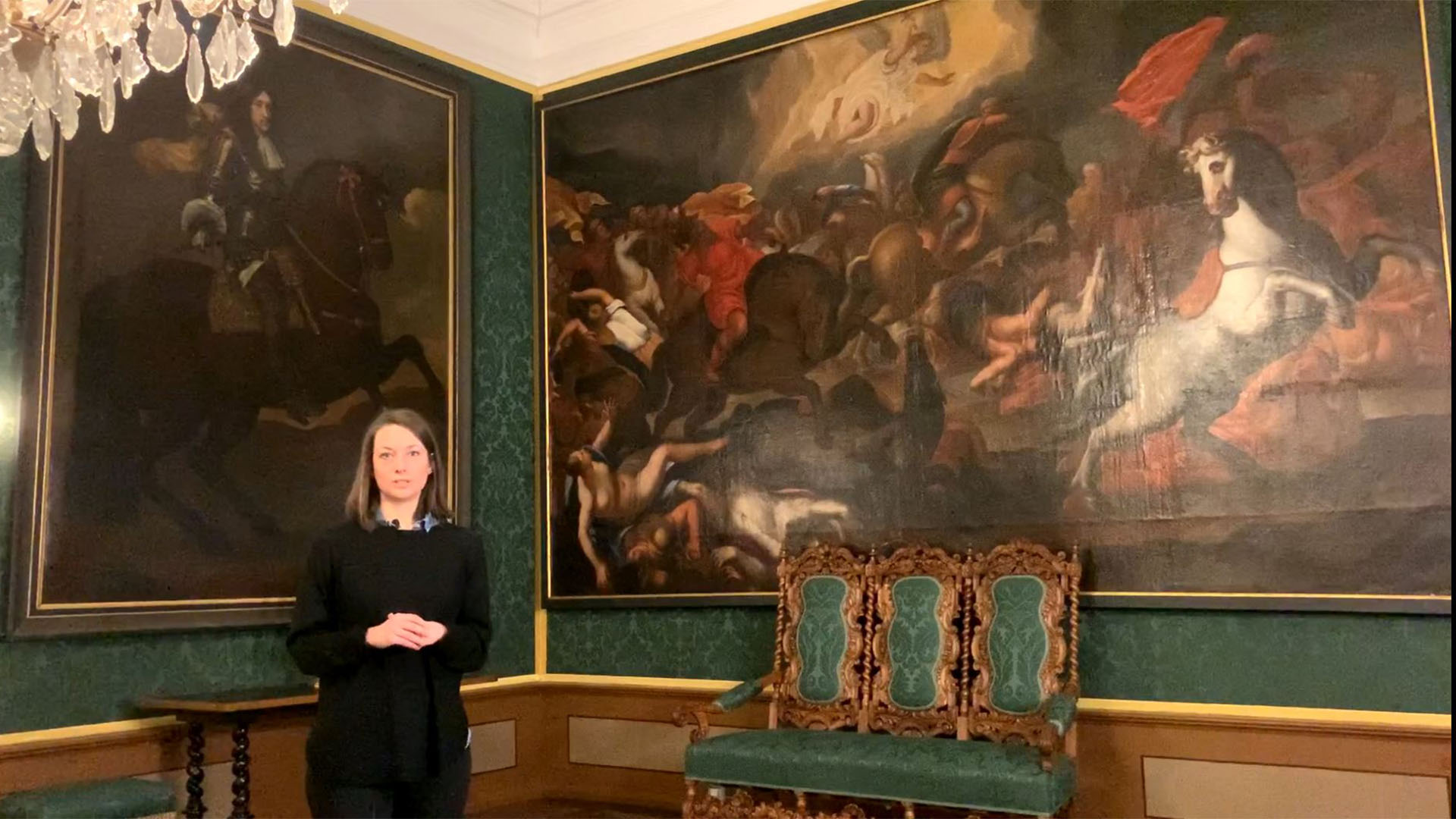 Stella Gilfert in einem Raum mit grüner Wandbespannung und einer prunkvollen Holzbank. Hinter ihr zwei großformatige Gemälde.