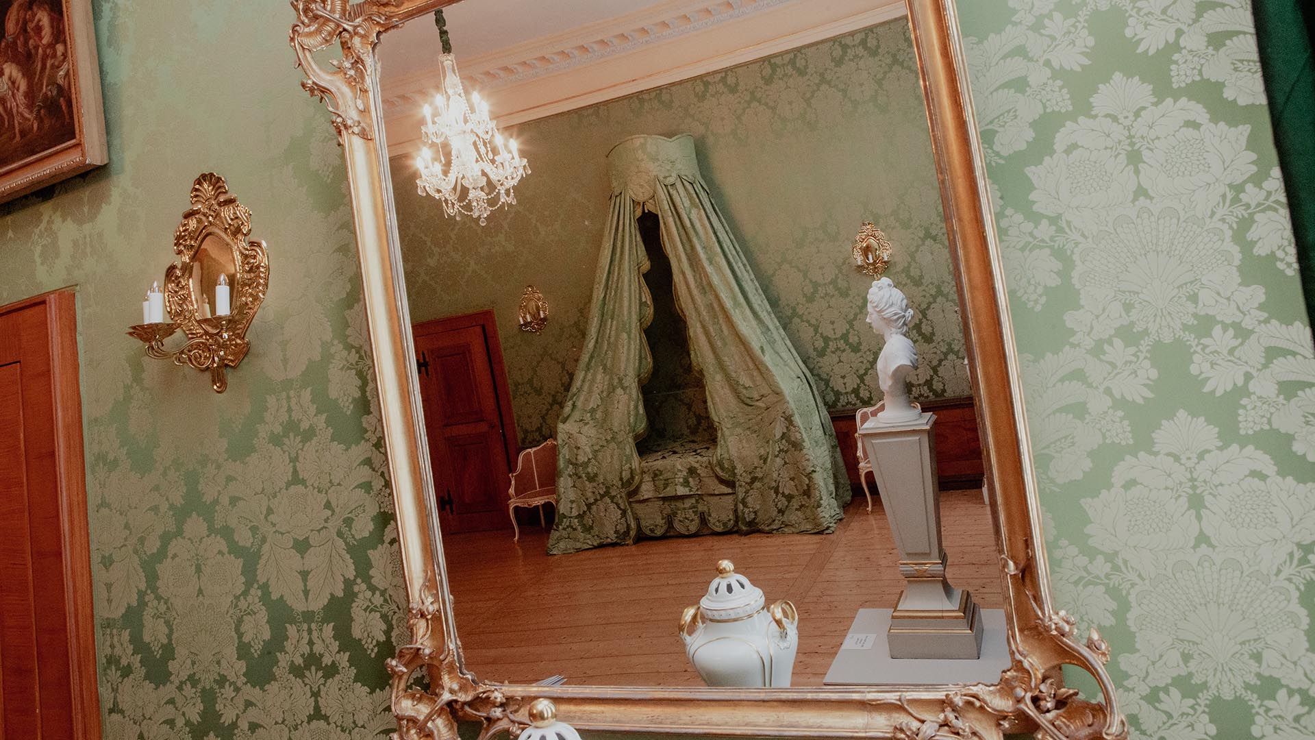 Blick in einen Wandspiegel, darin sieht man das Geburtszimmer der Herzogin Anna Amalia mit einem Himmelbet, einer Büste, Kronleuchter und kleinen Dekogegenständen