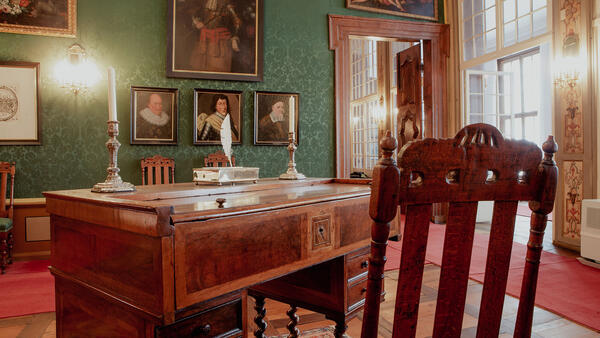 In einem Zimmer mit grünen Textiltapeten steht ein historischer Sekretär mit Kerzen und Federkiel. An der Wand hängen historische Porträts, davor stehen Stühle. Auf dem Parkettfußboden liegen Teppiche.