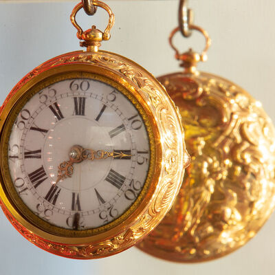Eine antike goldene Taschenuhr hängt so vor eienem Spiegel, dass man darin ihre kunstvoll verzierte Rückseite erkennen kann.