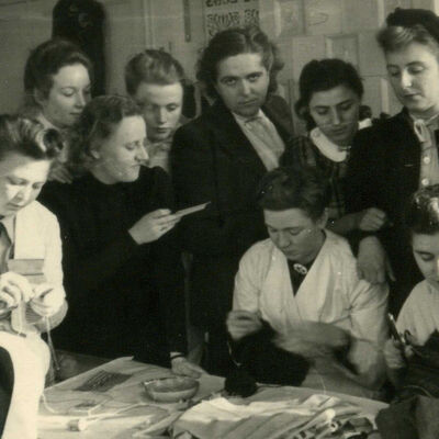 historisches Schwarzweißfoto: zehn Frauen sitzen und stehen um einen Tisch mit Stoffen, einige halten Stoffen in den Händen oder stricken.