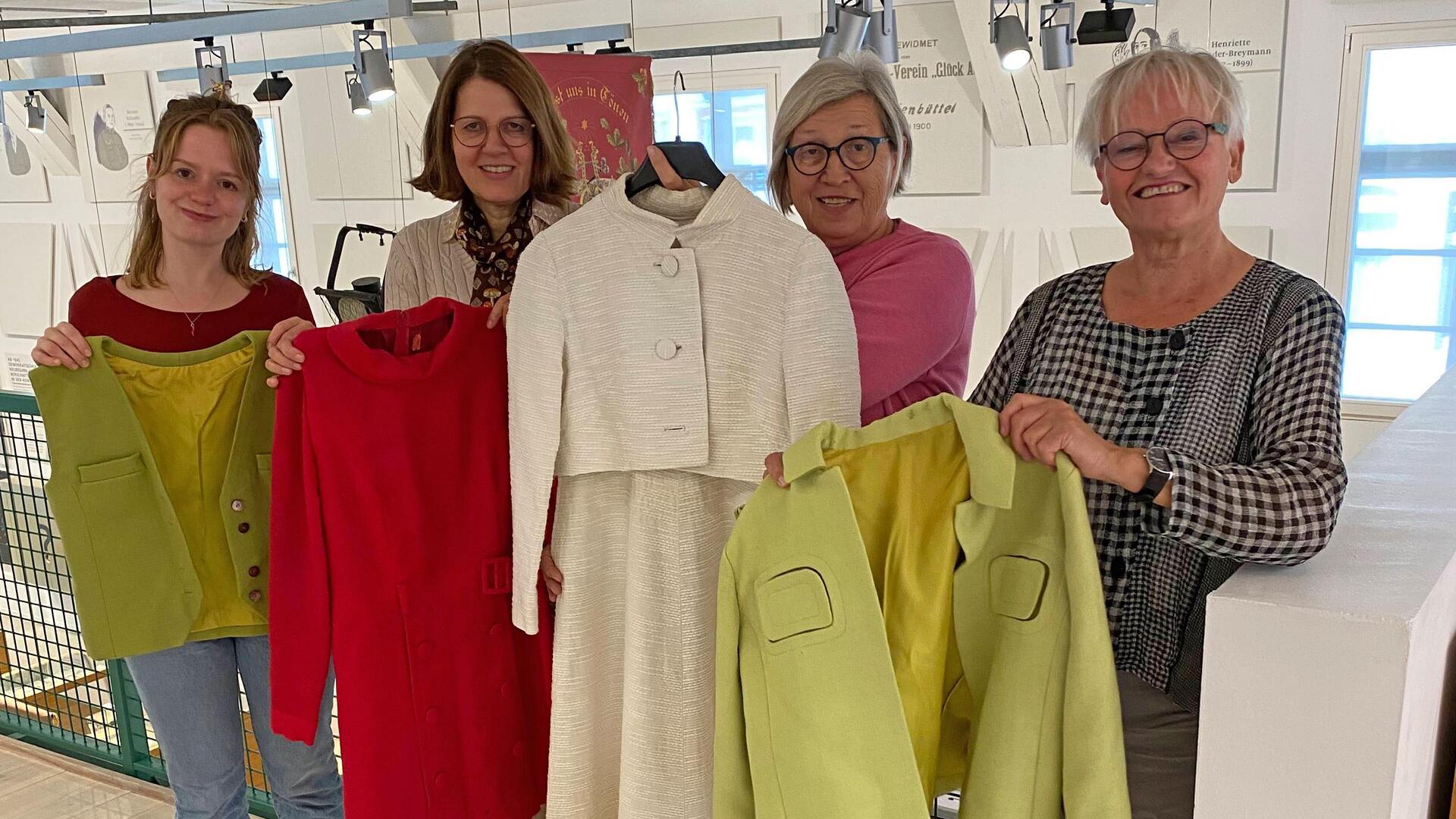 Vier Frauen stehen im Bürger Museum, jede hält ein Kleidungsstück wie eine Jacke oder Kostüm hoch.
