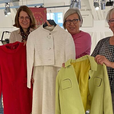 Vier Frauen stehen im Bürger Museum, jede hält ein Kleidungsstück wie eine Jacke oder Kostüm hoch.