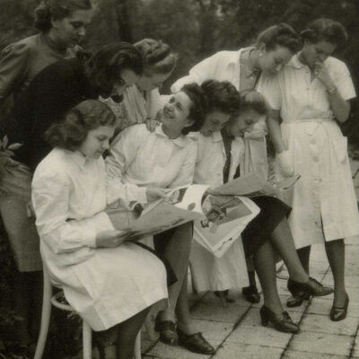 historisches Schwarzweißfoto: neuen Frauen, teilweise in weißen Kitteln, stehen und sitzen zusammen und schauen Modemagazine an.