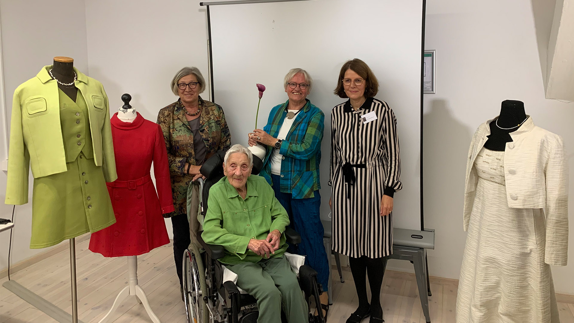 Das Foto zeigt Gabi Schrader, Ingrid Götze, Sieglinde Eberhard und Claudia Hagemann nach dem Vortrag über die Kleidermacherin Ruth Zimmermann im Bürger Museum. Sie stehen vor einer Leinwand, rechts und links von Ihnen befinden sich Kleider aus den 60er Jahren.
