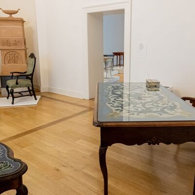 Ein Ausstellungsraum mit einem Schreibtisch mit buntem Perlenmotiv und Lehnstuhl, im Hintergrund ein Kamin mit Lehnstuhl und kleinem Tisch