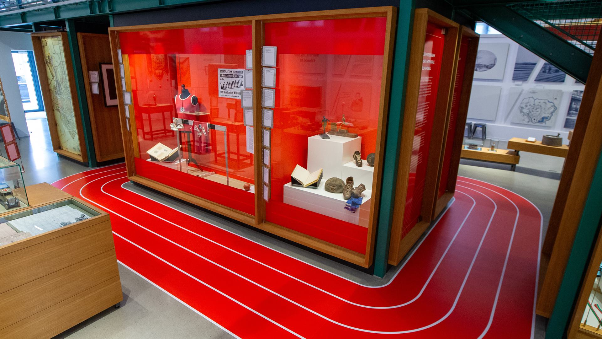 Eine Großraumvitrine mit Ausstellungsstücken ist mit rotem Hintergrund und Boden ausgestattet, um die Vitrinie läuft in rot eine nachgebildete Tartan-Laufbahn