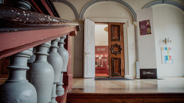 Das Foto zeigt den Eingangsbereich des Schloss Museums. Im Vordergrund zu sehen ist der letzte Absatz der Holztreppe und das hellgestrichene Geländer. Oben befindet sich die große, zweigeteilte Eingangstür und man blickt in den Empfangsraum mit Tresen.
