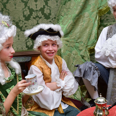 Das Foto zeigt drei Kinder um einen kleinen Holztisch. Das Mädchen ist als Prinzessin verkleidet und hält eine Porzellantasse in den Händen. Zwei Jungen im Prinzenkostüm, einer sitzend und einer stehend, schauen die Prinzessin an.